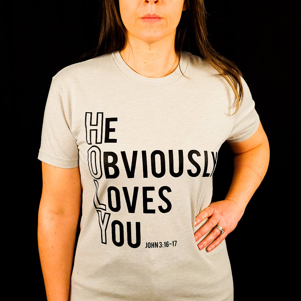 Beige t-shirt with the words He Obviously Loves You written downwards to show the word HOLY. The verse points to John 3:16-17