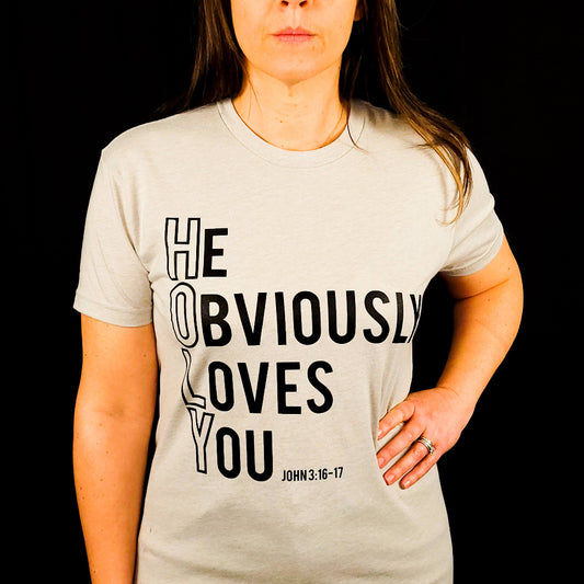 Beige t-shirt with the words He Obviously Loves You written downwards to show the word HOLY. The verse points to John 3:16-17