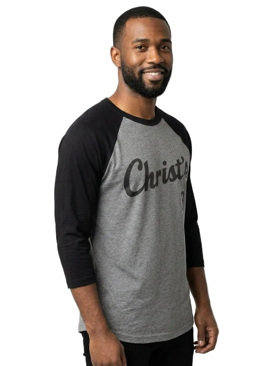 A man wearing a gray and black raglan baseball t-shirt with 'Christ's' printed in black script across the chest, photographed against a plain white studio background.