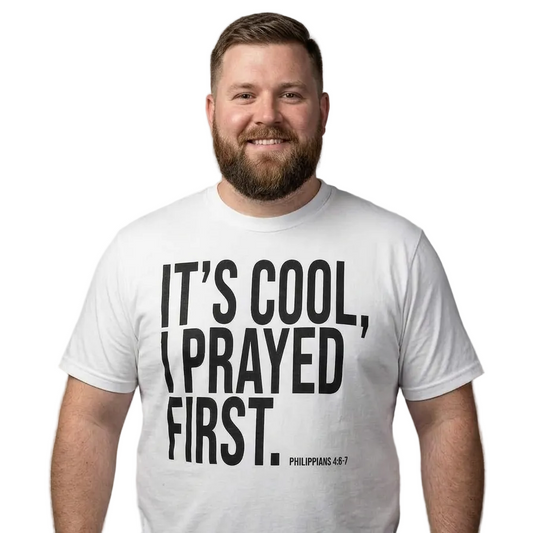 A smiling man with a beard wearing a white graphic t-shirt with the text 'IT'S COOL, I PRAYED FIRST. PHILIPPIANS 4:8?' printed in bold black letters, set against a plain gray studio background.