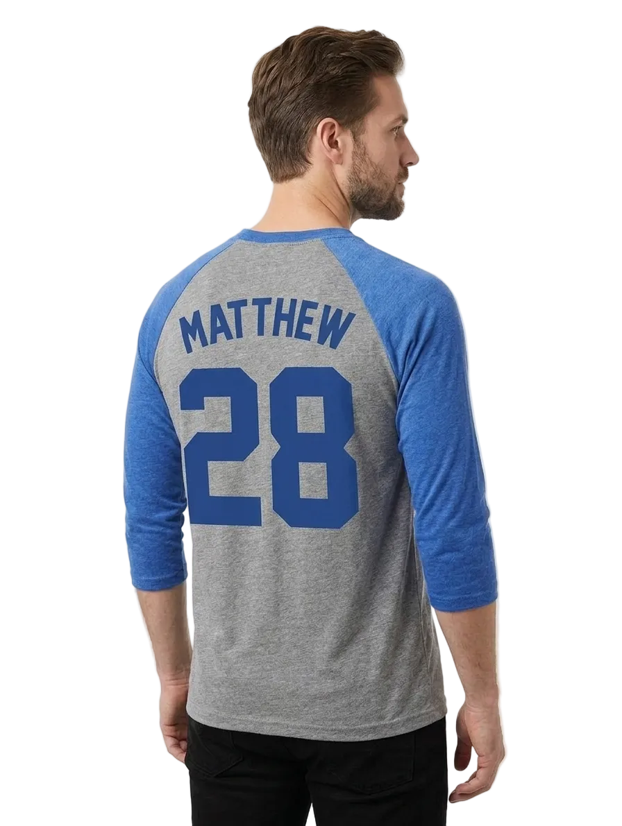 A man wearing a heather gray and blue raglan baseball t-shirt, viewed from the back. The shirt features the name 'MATTHEW' and number '28' printed in blue on the back.