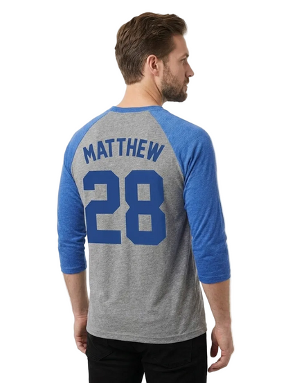 A man wearing a heather gray and blue raglan baseball t-shirt, viewed from the back. The shirt features the name 'MATTHEW' and number '28' printed in blue on the back.