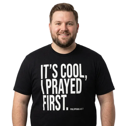 A man wearing a black short-sleeve t-shirt with the text 'IT'S COOL, I PRAYED FIRST. PHILIPPIANS 4:5-7' printed in bold white letters on the front, set against a plain white background.