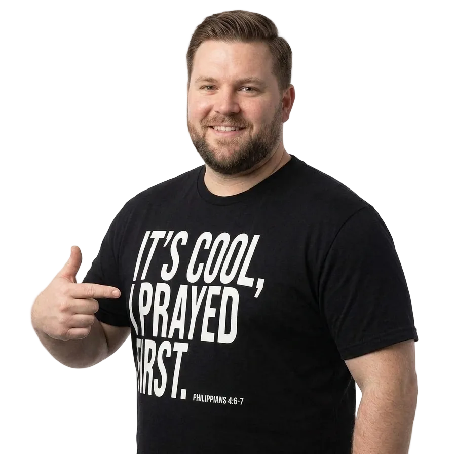 A smiling man wearing a black graphic t-shirt with the text 'IT'S COOL, I PRAYED FIRST. PHILIPPIANS 4:6-7' in white, pointing to the text with a thumbs-up gesture against a white studio background.