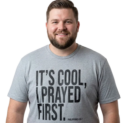 A smiling man with a beard wearing a heather gray t-shirt with the black printed text 'IT'S COOL, I PRAYED FIRST. PHILIPPIANS 4:6-7' on the front, set against a plain white background.