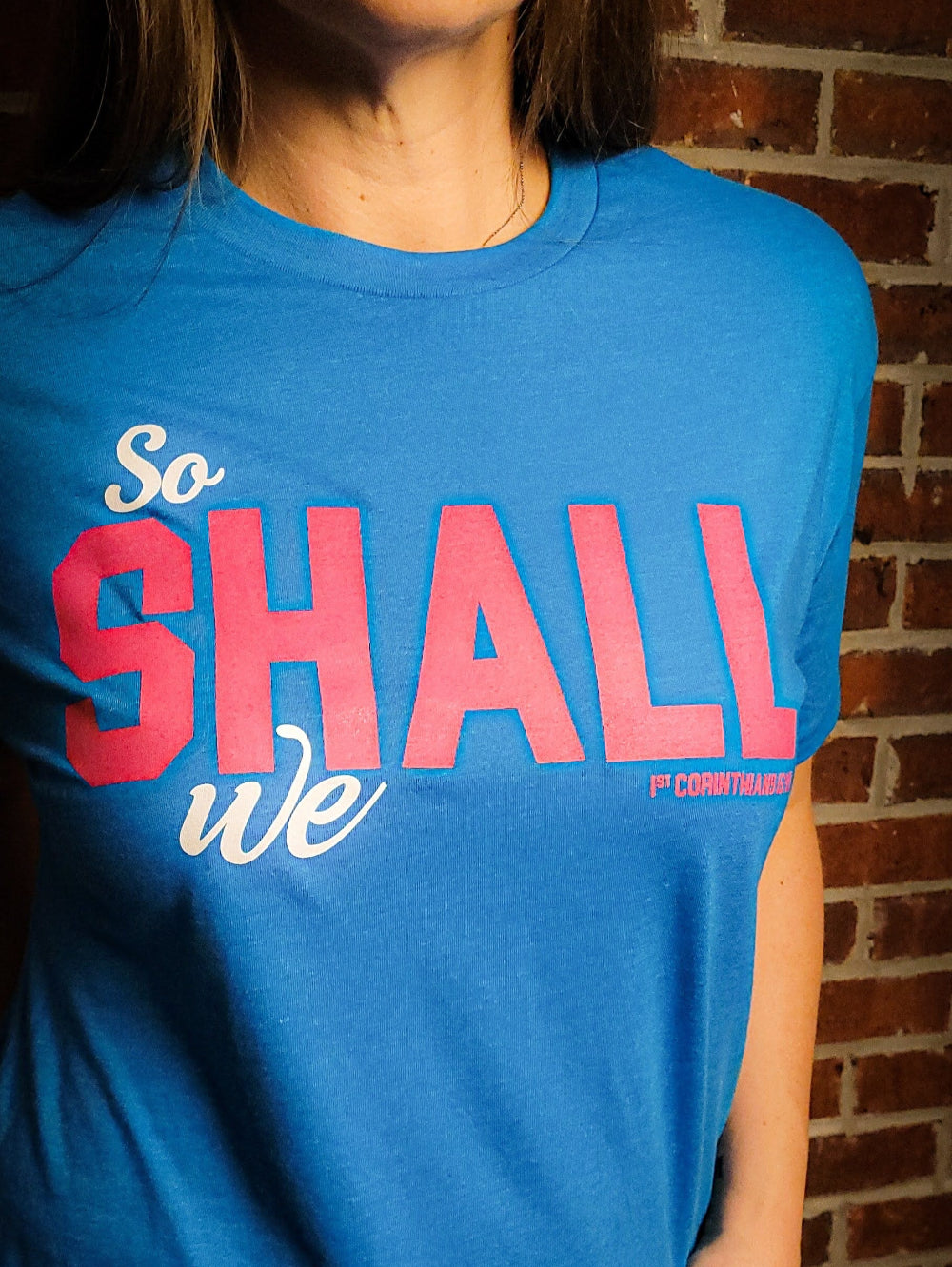 Teal blue shirt with the words So shall we across the front. The verse mentioned with 1 Corinthians 